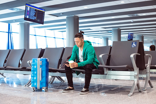 Young woman wearing green hoodie sitting in waiting room in the airport departure area and using phone, holding cup of coffee.