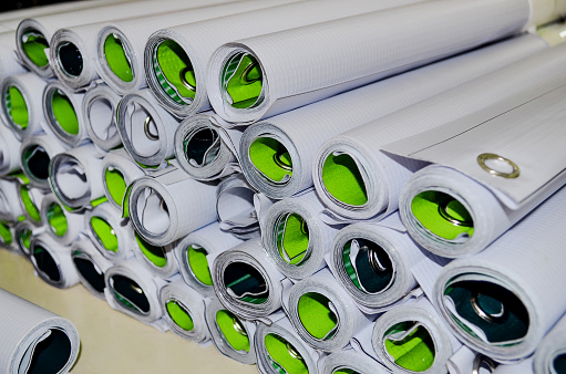 close up of a bunch of banners rolled up in a room in the printing industry. stacks of products are packed in the workspace. white tarpaulin printed in green.