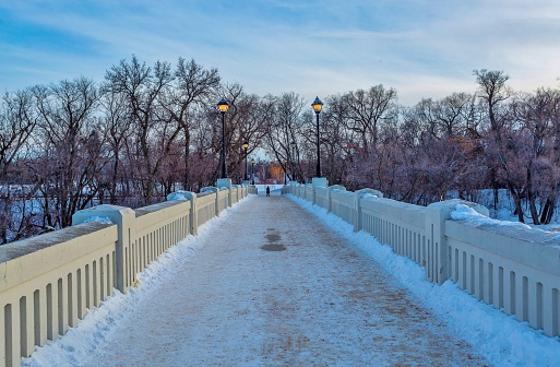 Dusk falls on the white concrete footbridge over the Assiniboine River to Assiniboine Park, Winnipeg, Manitoba, Canada.  Snow lies on the ground and the  sky is blue with fir trees in the background.