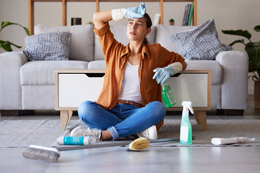 Tired woman, cleaning and sitting on living room floor with detergent, chemicals or sanitizer spray for hygiene at home. Female domestic worker exhausted from housekeeping, chores or clean house