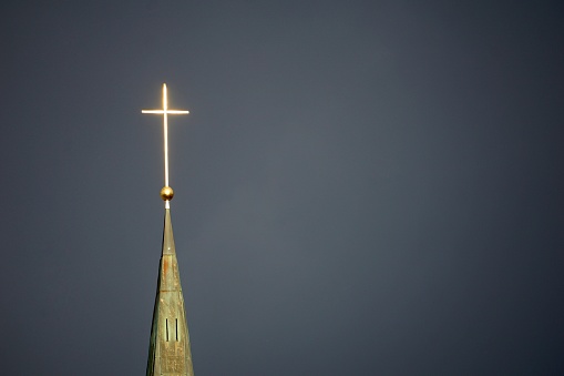 A shiny Catholic cross on the church in cloudy weather. A Catholic cross glitters in front of the sun against a cloudy black sky.