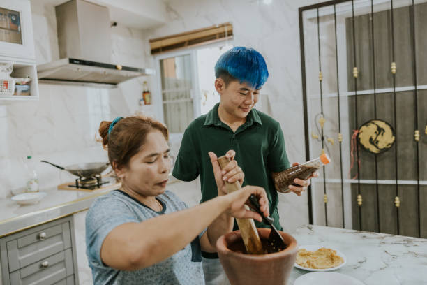 Real Thai LGBTQIA couple making lunch of a favorite Thai dish, spicy Som Tam with pickled fish. A non-binary person with an amputated right arm is picking up a bottle of pickled fish and giving to their girlfriend, who is a wheelchair user, as they make Som Tam together. transgender-athletes stock pictures, royalty-free photos & images