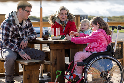 A family with two children sitting at a picnic table at an outdoor cafe area while enjoying a day out in Beadnell, North East England. They are enjoying refreshments while bonding together. The children are playing with pebbles on the table and the eldest daughter is a wheelchair user.