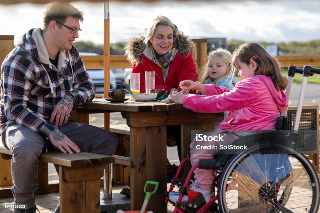 Simple Staycations A family with two children sitting at a picnic table at an outdoor cafe area while enjoying a day out in Beadnell, North East England. They are enjoying refreshments while bonding together. The children are playing with pebbles on the table and the eldest daughter is a wheelchair user. Family Stock Photo