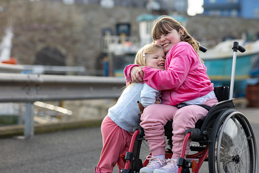 Two girls enjoying a day out together in Beadnell, North East England. They are embracing each other next to the harbour while smiling. The eldest girl is a wheelchair user.