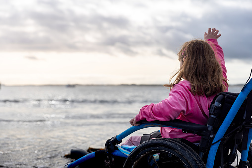 A young girl in the sea waving to a boat in the sea at Beadnell beach, North East England. She is a wheelchair user.