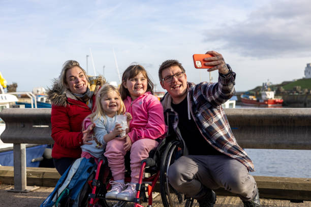 Making Great Memories A family with two children enjoying a day out together in Beadnell, North East England. They are next to the harbour and taking a selfie on a mobile phone together. The eldest daughter is a wheelchair user. special needs child pictures stock pictures, royalty-free photos & images
