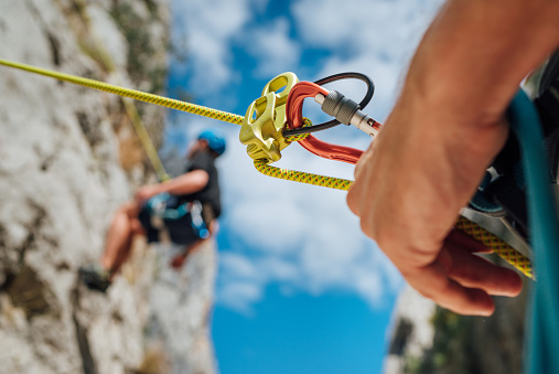 Belay device close-up shot with a boy on the cliff climbing wall. He hanging on a rope in a climbing harness and his partner belaying him on the ground. Active people and sports concept image