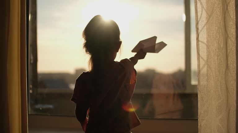 A child plays with a paper plane near the window of the house during sunset.Authentic scene.