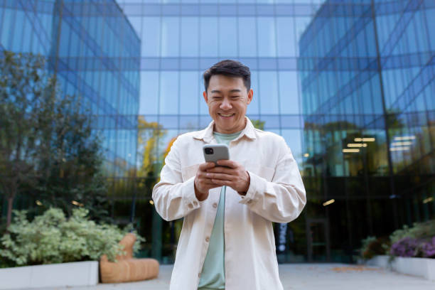 istock Asian man using phone outside office building, freelancer businessman typing message and smiling, programmer in casual clothes browsing online web pages from app 1473735691