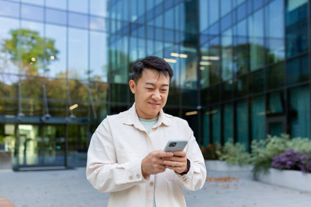 istock Asian man using phone outside office building, freelancer businessman typing message and smiling, programmer in casual clothes browsing online web pages from app 1473735697