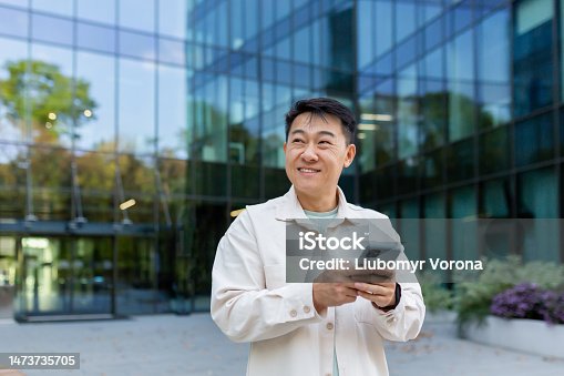 istock Smiling asian man in casual shirt walking in city using app on smartphone, man looking to side on background of office building from outside 1473735705