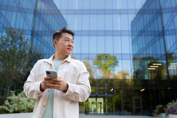 istock Smiling asian man in casual shirt walking in city using app on smartphone, man looking to side on background of office building from outside 1473735708