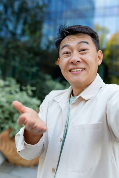 istock Asian programmer talking on video call using app on phone, view camera man waving and talking cheerfully, outside office building on break. 1473735715