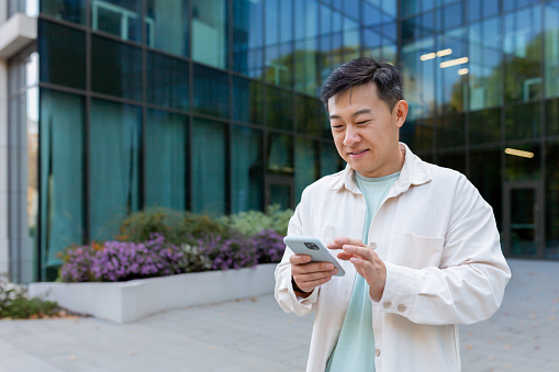 Asian smiling and happy watching online video on phone outside office, man typing message in casual clothes using smartphone.