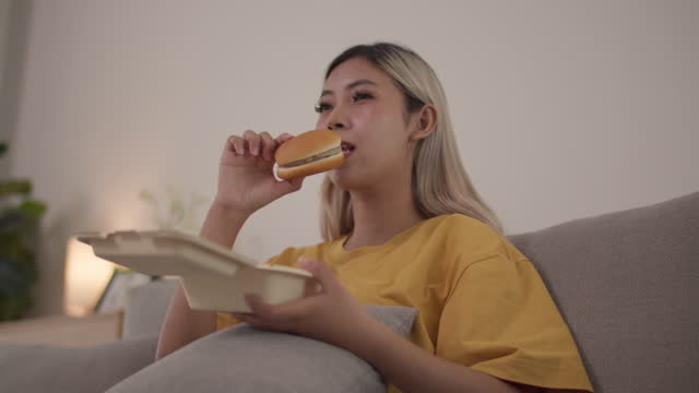 Asian woman eating burger in the living room at home.