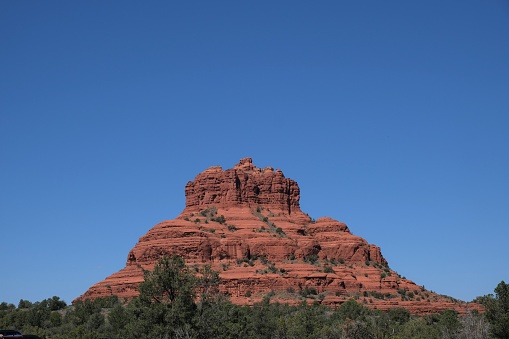 Bell rock formation which is red sandstone at different times of day and from different distances against a blue sky surrounded by scrub brush and local trees. Located in Sedona, AZ is perfect area for outdoor enthusiasts and hikers.