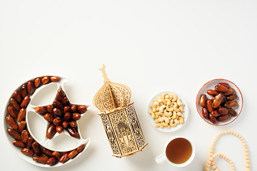 Muslim holiday Ramadan Kareem concept. Flat lay dried dates in Islamic star and crescent plate, nuts, oriental lantern, cup of tea, rosary on white background.
