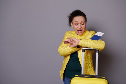 Multi-ethnic woman in bright yellow jacket, checks time on her wrist watch, expressing disappointment and worry, feeling anxious being unpunctual at flight, posing with open mouth on gray background