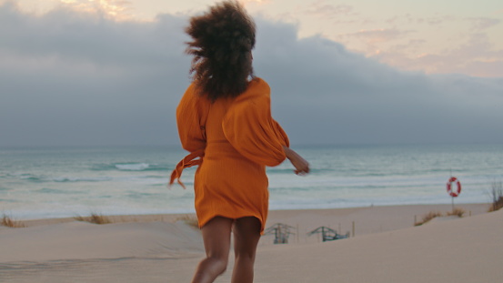 Cheerful woman escaping on sand beach in front gray cloudy sky. Happy african american girl wearing orange dress running on seashore smiling playfully summer evening. Curly brunette have fun on nature