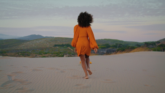 Back view of woman jogging on desert wearing orange dress at summer twilight. Unknown curly girl crouching on sand. Active african american brunette relax on beautiful dunes at gloomy evening.