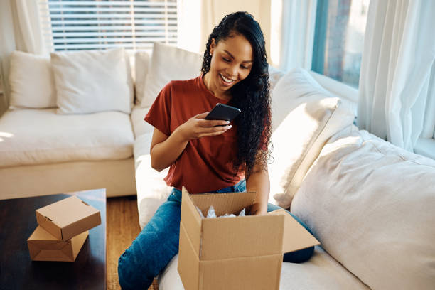 istock Happy black woman using cell phone while unpacking delivery box at home. 1473763318