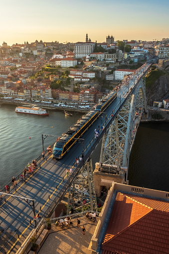 View from the Serra do Pilar viewpoint in Vila Nova de Gaia, over the upper deck of the D. Luis bridge, with the metro and tourists crossing the bridge at the end of the afternoon in summer. The city of Porto with the Ribeira, the houses and the Cathedral serve as a backdrop.