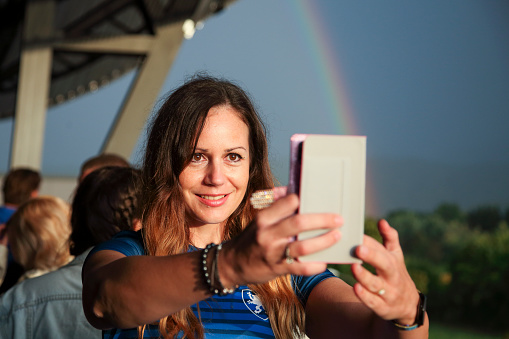 Female fan on the outside of a stadium taking selfie with smart phone with rainbow in the background. Positive emotion. All logos are property released.