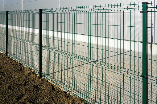 A new green metal grid fence around a new industrial site, grass soil on one side and a paved lane on the other