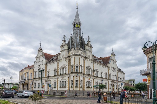 Caransebes, Romania - July 6, 2022: Beautiful building of town hall, street with cars and some people walking around. Overcast sky.  It is located in Banat region in southwestern Romania