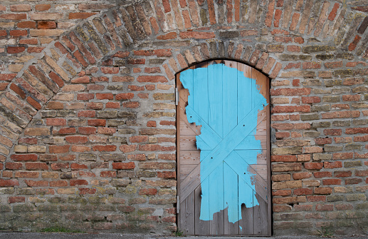 Background of a brick wall with a door in it. A round arch is above the door, made of stones. The wooden door is closed and smeared with blue paint.