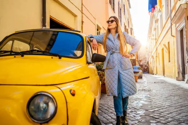 Woman leaning against vintage yellow Fiat 500 parked along an alley in Rome, Italy