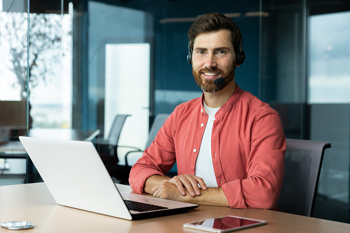 Portrait of mature businessman in red shirt inside office, man with beard and video call headset smiling and looking at camera, online customer support worker with laptop.