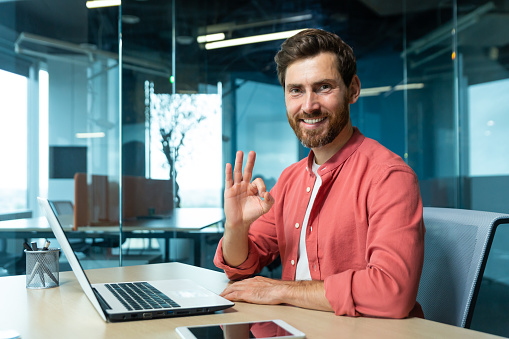 Portrait of a successful young man programmer, designer, freelancer in a red shirt sitting and working in the office at a laptop. He looks at the camera smiling. shows ok with hand.