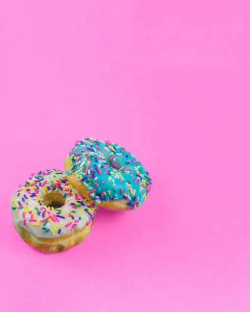 Two donuts with blue and white icing and colorful sprinkles on a plain bright pink background with room for copy space.