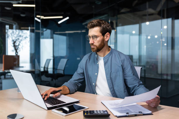 ein mann finanzier, wirtschaftsprüfer sitzt im büro. er hält dokumente, prüft konten. typen auf einem laptop - buchprüfung stock-fotos und bilder