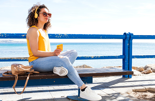 Curly lady in sunglasses listening to music in headphones from cellphone while sitting on wooden bench on embankment in daylight
