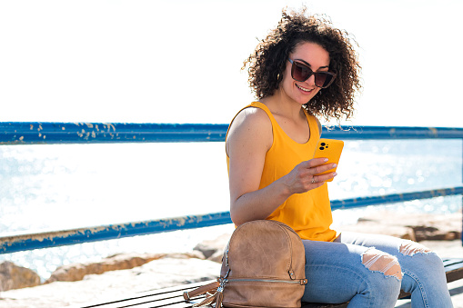 Positive young curly haired female in casual clothes and sunglasses sitting on bench with backpack and smiling while browsing smartphone against sea during summer vacation