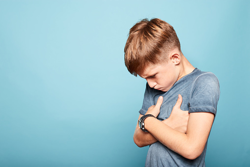 Frustrated caucasian young boy with arms crossed standing sideways, isolated on blue background. Copy space.