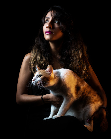 A woman sitting in a dark room with a white and yellow cat on her lap, illuminated by two bright lights on either side