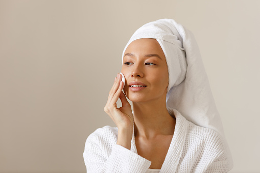 Young woman in white robe and towel on her head wipes the skin of her face with cotton pads. Cleansing and healthy body care after shower. Close-up of pretty girl on isolated background