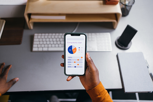 A from above of an unrecognizable African-American entrepreneur analyzing some statistics using his smartphone. He is sitting at his office desk. (close up view)