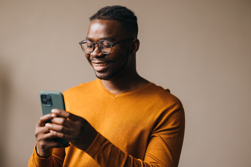 A smiling mixed-race African-American entrepreneur watching something on his smartphone while standing against white background. (studio shot, copy space)