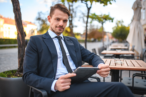 Medium shot of young business man working on his digital tablet, while sitting at a table of outside cafe, looking at camera.
