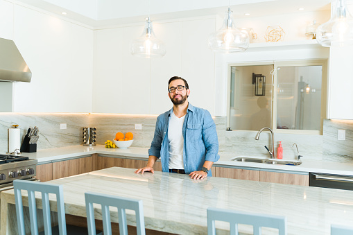 Happy attractive man in his 30s smiling making eye contact while enjoying his luxury granite white kitchen