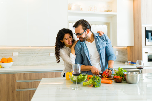 Smiling couple in love hugging while cooking a healthy dinner together on a white granite kitchen island
