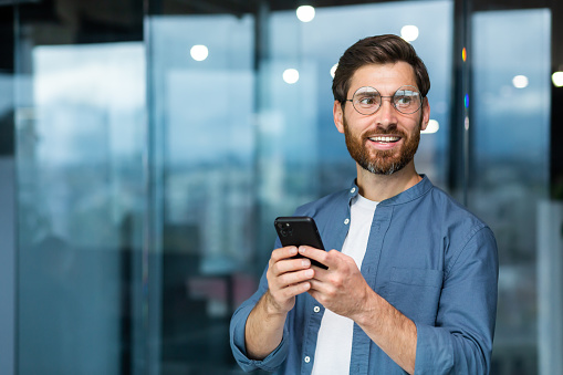 Portrait of a young handsome male student in glasses standing in the campus, office. He holds the phone in his hands, smiles, looks to the side.
