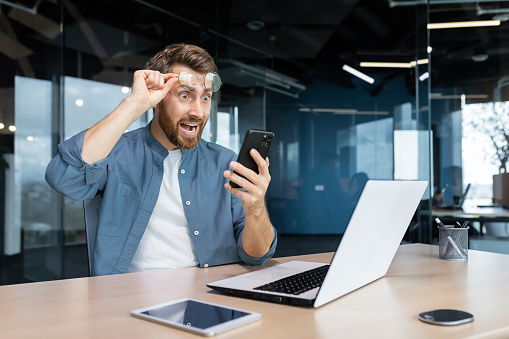 Mature man shocked by bad news and online notification received on phone, businessman at work frustrated and shouting at smartphone screen, worker at workplace sitting with laptop.