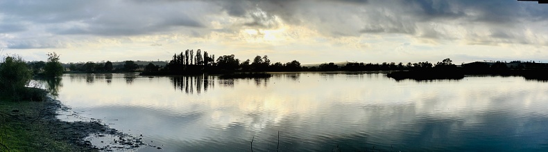 Horizontal panoramic view of trees, in silhouette and a stormy sky reflecting on the waters of the wildlife refuge at Dangar’s Lagoon, a habitat for migratory and endemic bird species near Armidale, New England high country, NSW.