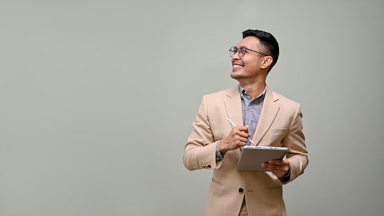 Smiling and handsome millennial Asian businessman in a formal business suit and glasses, looking aside at the empty space, holding a tablet, stands against a green studio background.